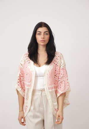 Donna con capelli lunghi e scuri indossa un top bianco, pantaloni beige e uno scialle lavorato all'uncinetto rosa e bianco, in piedi davanti a uno sfondo chiaro uniforme.