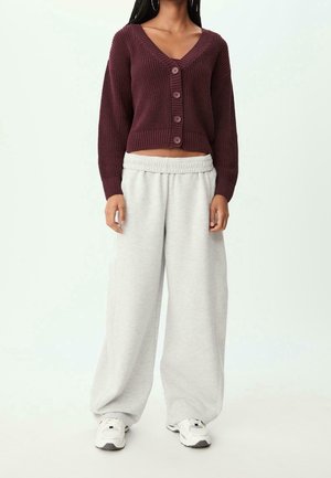 Femme portant un cardigan court bordeaux boutonné, un pantalon large en molleton gris clair et des baskets blanches, debout devant un fond uni.