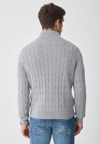 Maglione grigio a maglia con colletto alto e orlo a coste. Design a trama e maniche lunghe, indossato con jeans azzurri chiari.