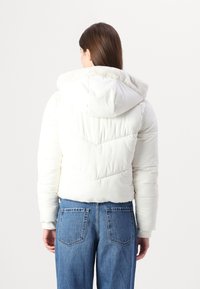 White puffer jacket with a textured fur-lined hood, featuring chevron stitching and elastic cuffs, paired with blue high-waisted jeans.