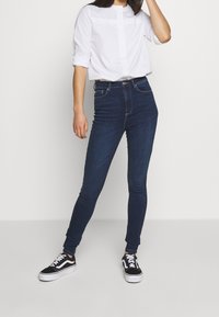 Jeans de tiro alto en azul oscuro, hechos de denim, con un ajuste slim y detalles de costura sutiles. Combinado con una camisa blanca de botones.