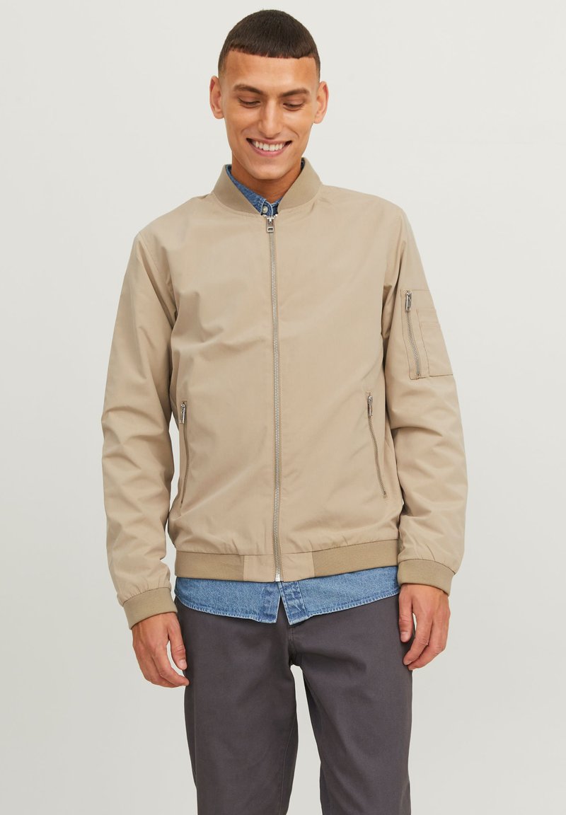 Veste bomber beige avec une fermeture éclair sur le devant, des poches latérales et des poignets élastiques. Le tissu semble lisse avec une finition légèrement brillante.