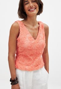Coral sleeveless top with a floral texture, featuring a V-neckline and layered rose patterns. Worn with white linen pants and bangle bracelets.