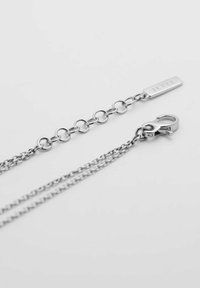 Silverfärgad kedja med alternerande släta och texturerade länkar, med en kräftklämma och en rektangulär tagg graverad med "JETTE."