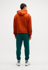 Felpa arancione con cappuccio e vestibilità comoda, abbinata a pantaloni della tuta verdi. I pantaloni presentano polsini a coste; entrambi realizzati in morbido misto cotone.