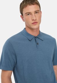 Młody mężczyzna z krótkimi włosami, ubrany w niebieską koszulę polo z krótkim rękawem i dwoma guzikami, na jednolitym białym tle.
