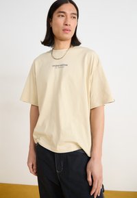 Beige bomull T-skjorte med korte ermer, med et sentralt "STUDIO EDITION" grafikk. Brukt med svarte bukser, som viser en avslappet passform.