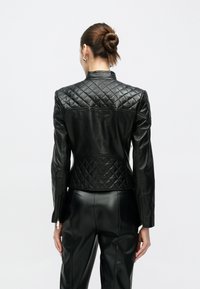 Μαύρο δερμάτινο μπουφάν με καπιτονέ υφή στο πάνω μέρος της πλάτης. Κομψή εφαρμογή με μακριά μανίκια και ψηλό γιακά, σχεδιασμένο με ελάχιστο μεταλλικό εξοπλισμό.