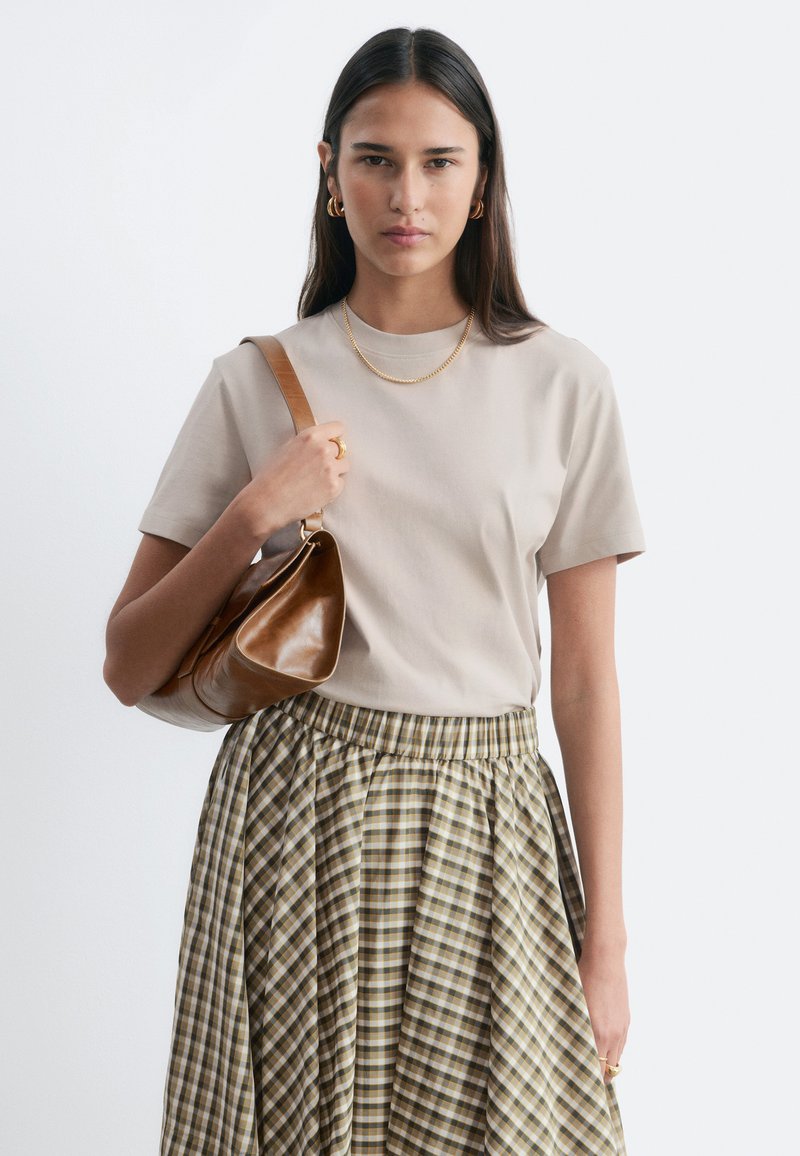 Femme aux longs cheveux foncés portant un t-shirt beige et une jupe à carreaux, tenant un sac à bandoulière en cuir marron, debout devant un fond uni.