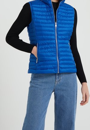 Persona que lleva un chaleco acolchado azul con cremallera sobre una camiseta negra de manga larga y azul de denim, con una mano en un bolsillo del chaleco.