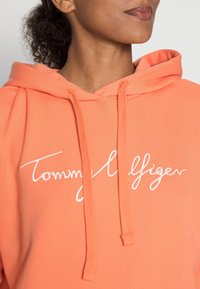 Felpa con cappuccio di un vivace color corallo, con logo "Tommy Hilfiger" ricamato in bianco. Realizzata in tessuto morbido, presenta cordini e una vestibilità comoda.