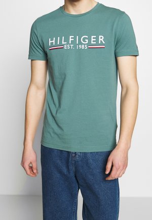 Un homme portant un t-shirt à manches courtes teal avec le texte "HILFIGER EST. 1985" sur la poitrine et un jean bleu, se tenant contre un fond uni.