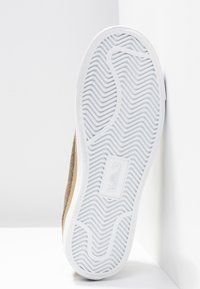 Semelle de basket blanche avec un motif ondulé et une zone rectangulaire pour le logo, positionnée à la verticale contre des murs d’angle blancs.