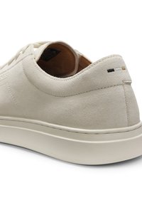 Μπεζ σουέτ αθλητικό παπούτσι με λευκή καουτσούκ σόλα, χαμηλή γλώσσα αστραγάλου και τρεις μικρές ραμμένες χρωματικές ρίγες σε μαύρο, λευκό και ταμπ στο πίσω μέρος.