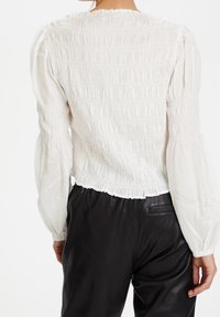 Blusa bianca con arricciature, maniche a sbuffo e orlo a volant, abbinata a pantaloni di pelle neri. Tessuto morbido e strutturato.