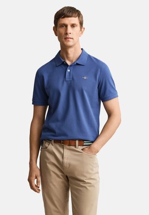 Hombre que lleva una camiseta polo azul de manga corta y pantalones beige, de pie con una mano en el bolsillo frente a un fondo liso.
