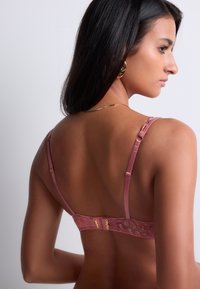 Soutien-gorge en dentelle rose avec des bretelles réglables, une fermeture à crochet et à œillet, et une finition satinée lisse, ornée de détails floraux le long de la bande.