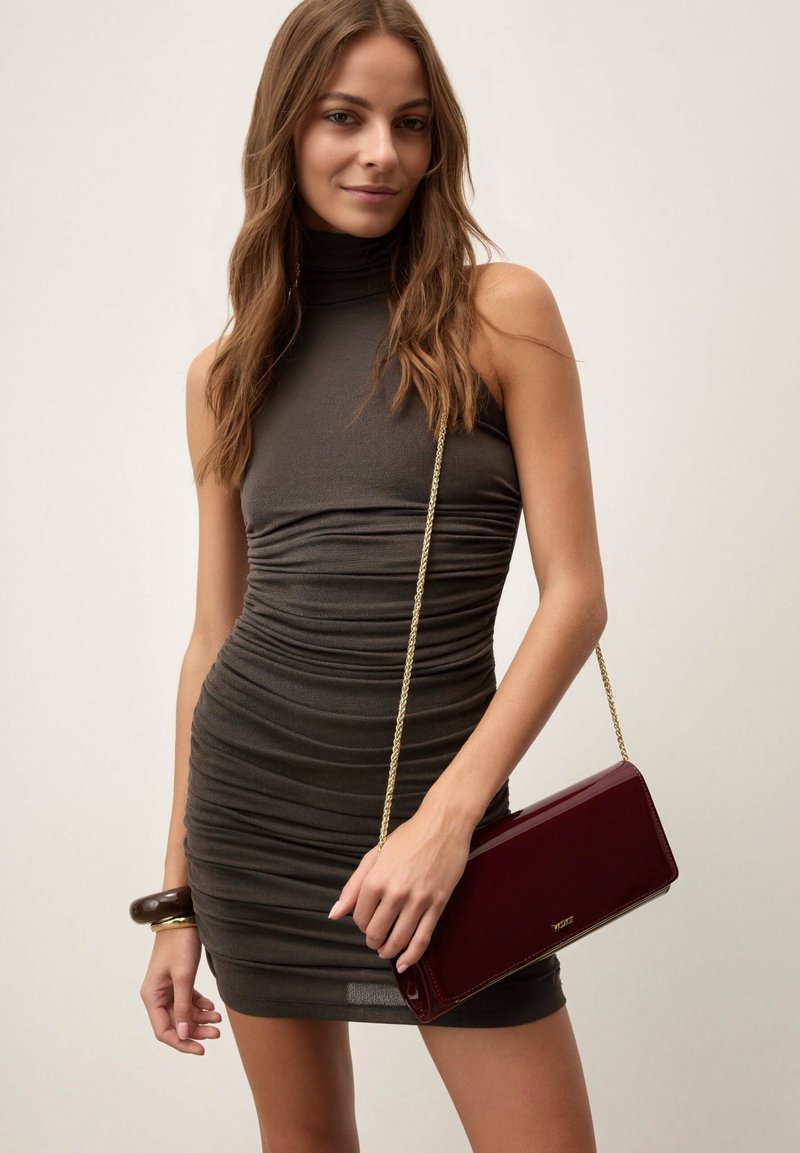 Nainen, joka pukeutuu hihattomaan, ryppyiseen tummanharmaaseen mekkoon ja kantaa olalla viininpunaista laukkua, jossa on kultainen ketjuhihna.