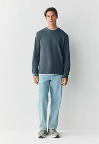 Grå, struktureret langærmet skjorte med hvid krave, kombineret med lyseblå jeans og beige sneakers. Modellen står lige op ad en neutral baggrund.