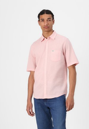 Camicia a maniche corte, di colore rosa chiaro, con bottoni e realizzata in un tessuto texturizzato. Presenta una tasca sul petto con un piccolo logo. Indossata con jeans blu.