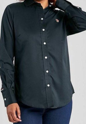 Persona que lleva una camisa negra de manga larga con botones y un pequeño logo bordado en el pecho, combinada con vaqueros azules.