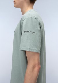 T-shirt di cotone verde chiaro con maniche corte. Presenta un logo stampato "Napapijri Geographic" e "SINCE 1987" sulla manica sinistra.