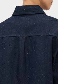 Donkerdenimjack met een gestructureerd oppervlak en kleine, glanzende accenten die gelijkmatig verdeeld zijn. Klassieke kraag en rugdetails.