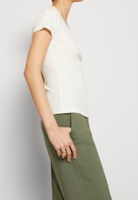 Valkoinen lyhythihainen toppi V-kauluksella, jonka kangas on ribbineulosta. Yhdistetty vihreisiin housuihin, jotka ovat rentoa mallia ja sileäpintaiset.