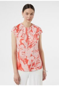 Koronkowa bluzka w kolorze koralowym z krótkimi rękawami, ozdobiona białym nadrukiem roślinnym, z plisowanym dekoltem i gładką fakturą. Połączona z białymi spodniami.