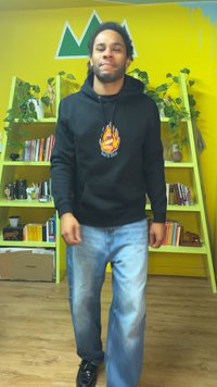 Sweat à capuche noir avec un motif de flammes, jean bleu usé et chaussures noires. Debout dans une pièce colorée avec des étagères vertes et des plantes.