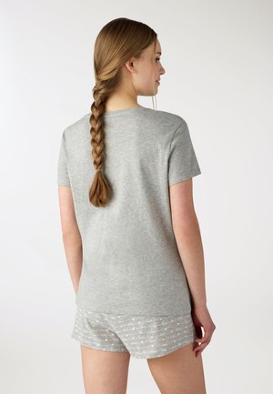 Donna con lunghi capelli intrecciati che indossa una maglietta grigia a maniche corte e pantaloncini grigi chiari con motivo, in piedi leggermente girata di lato.