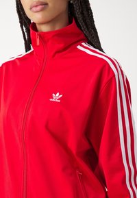 Czerwona kurtka zapinana na suwak, wykonana z gładkiej tkaniny. Posiada wysoki kołnierz, białe trzy paski na rękawach oraz małe białe logo na piersi.