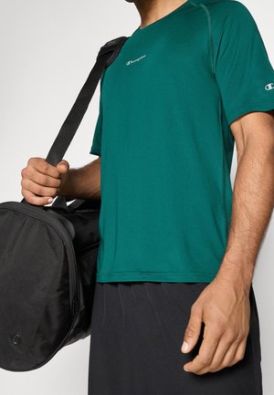 Person iført en grøn Champion t-shirt, der holder en sort duffelbag over den ene skulder mod en ensfarvet baggrund.