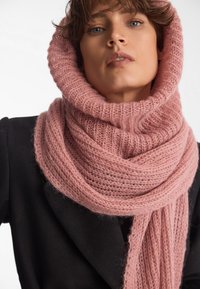 Miękki, różowy, dziany szalik o grubym ribbowym wzorze. Ma długie końce opadające w dół, noszony na czarnym płaszczu. Uzupełnia całość stylizacji.