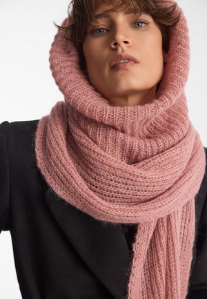 Miękki, różowy, dziany szalik o grubym ribbowym wzorze. Ma długie końce opadające w dół, noszony na czarnym płaszczu. Uzupełnia całość stylizacji.