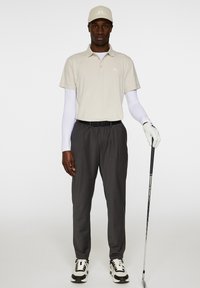 Beige pikétröja med logotyp, lager på lager under en vit långärmad skjorta, grå byxor, svart bälte, vit golfhandske och svart/vita sneakers.