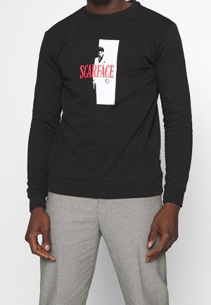 Sudadera negra con cuello redondo, que presenta un diseño gráfico en blanco y rojo de "Scarface" en la parte delantera. Fabricada en tejido suave con puños de canalé.