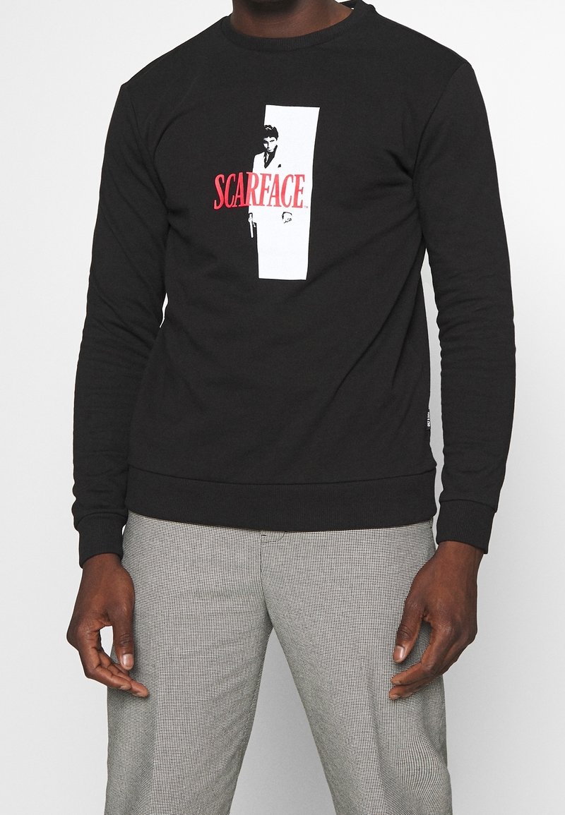 Sudadera negra con cuello redondo, que presenta un diseño gráfico en blanco y rojo de "Scarface" en la parte delantera. Fabricada en tejido suave con puños de canalé.