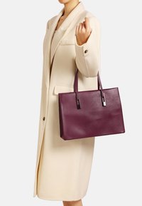 Fioletowa torba tote ze sztywną konstrukcją, błyszczącymi srebrnymi akcentami i dwoma uchwytami, trzymana przez osobę w kremowym płaszczu.