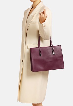Borsa tote in pelle viola con un design strutturato, rifiniture argentate lucide e due manici, tenuta da una persona in un cappotto crema.