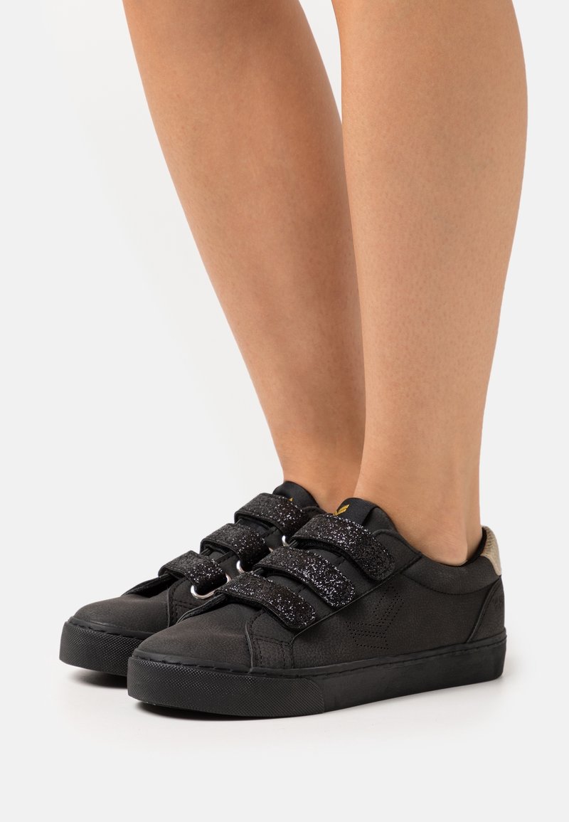 Baskets noires avec des brides Velcro noires pailletées, bout rond et matière texturée. Semelle noire unie avec un petit accent beige au talon.