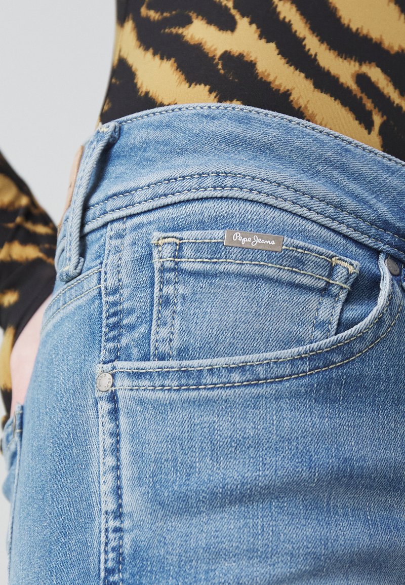 Jeans en denim bleu clair avec une étiquette emblématique près de la poche arrière, présentant une couture classique et une coupe épurée. Tissu texturé visible.