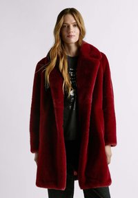 Cappotto di pelliccia rossa con colletto a lancia, caratterizzato da una texture morbida e una vestibilità comoda. Indossato sopra una maglietta nera con stampa grafica e pantaloni scuri.