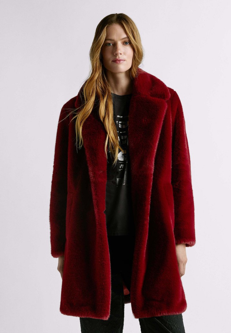 Cappotto di pelliccia rossa con colletto a lancia, caratterizzato da una texture morbida e una vestibilità comoda. Indossato sopra una maglietta nera con stampa grafica e pantaloni scuri.