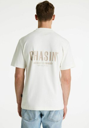 Muškarac odjeven u bijelu majicu s tekstom "CHASIN' AMS BASED - C.COMMUNITY" na leđima, u kombinaciji sa svijetloplavim trapericama, okrenut leđima.