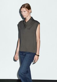 Top bez rękawów z kapturem w ciemnym oliwkowym kolorze, w połączeniu z niebieskimi spodniami prostymi. Gładka tekstura, minimalistyczny design i brak widocznych wzorów lub akcentów.