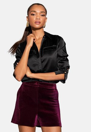 Black satin shirt with rolled sleeves and two front pockets, paired with burgundy velvety shorts featuring a button detail at the waist.