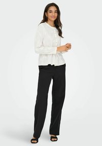 Chemisier blanc à manches longues avec un col rond, fermetures à boutons et un motif floral subtil. Associé à un pantalon noir à jambe droite.