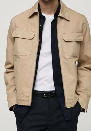 Giacca beige con chiusura a zip e tasche a patta indossata sopra una camicia nera con bottoni e una T-shirt bianca, abbinata a pantaloni neri e cintura.