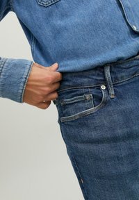 Sötétkék farmernadrág karcsúsított fazonnal, öt zsebbel, kontrasztos öltésekkel és gombos záródással. Lágy textúra látható.